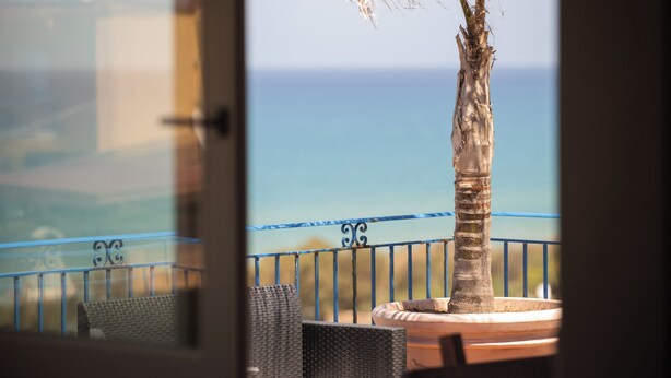 A balcony with wicker furniture and a palm tree, overlooking a coastal view.