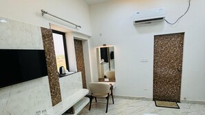 A hotel room with a bed, a TV mounted on a marble wall, a chair, a mirror, and an air conditioning unit. - Hotel Shri Tulsi Residency (Phalodi)