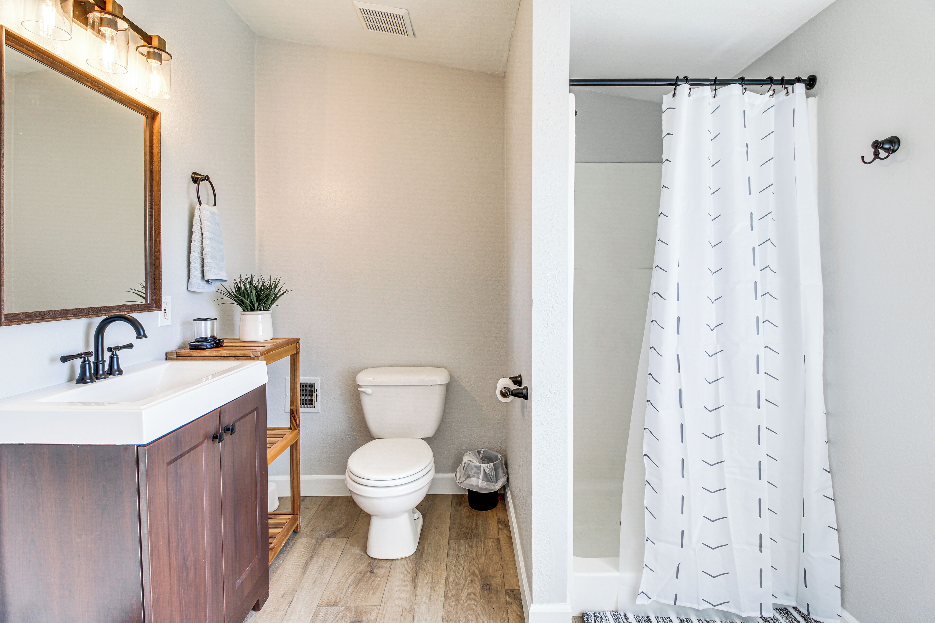 Un baño con tocador de madera, inodoro blanco, ducha con cortina estampada y espejo con iluminación.