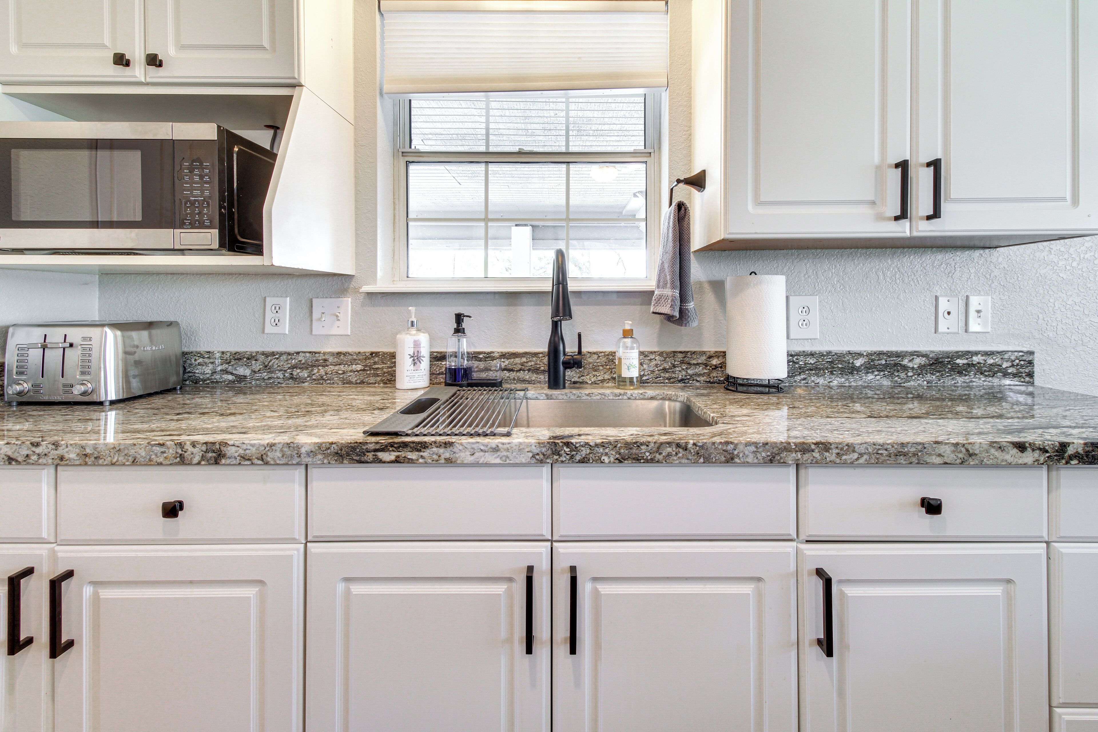 A kitchen with white cabinets, a granite countertop, a microwave, a toaster, and a window with blinds.