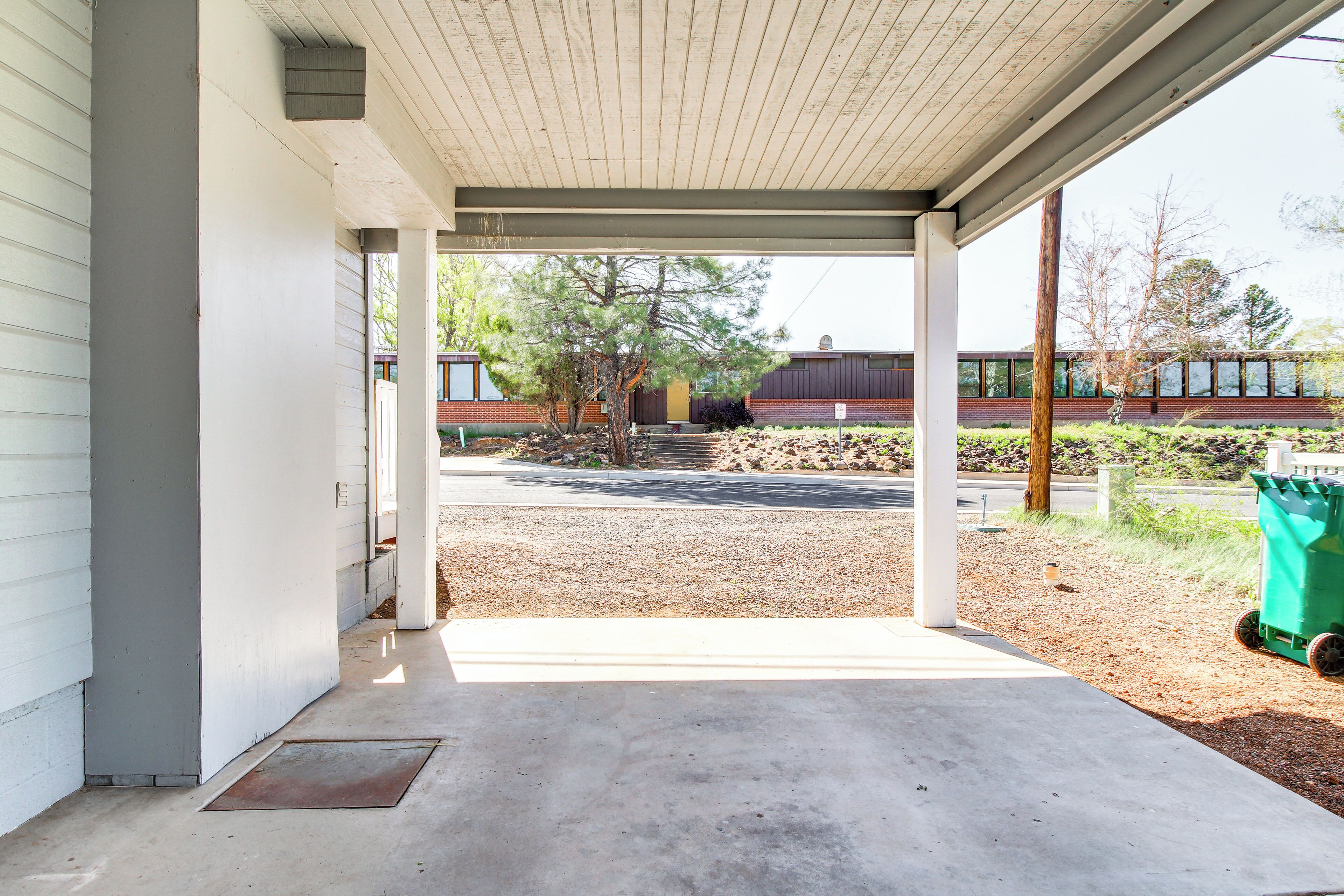 A covered outdoor area with a concrete floor, a green trash bin, and a building in the background.