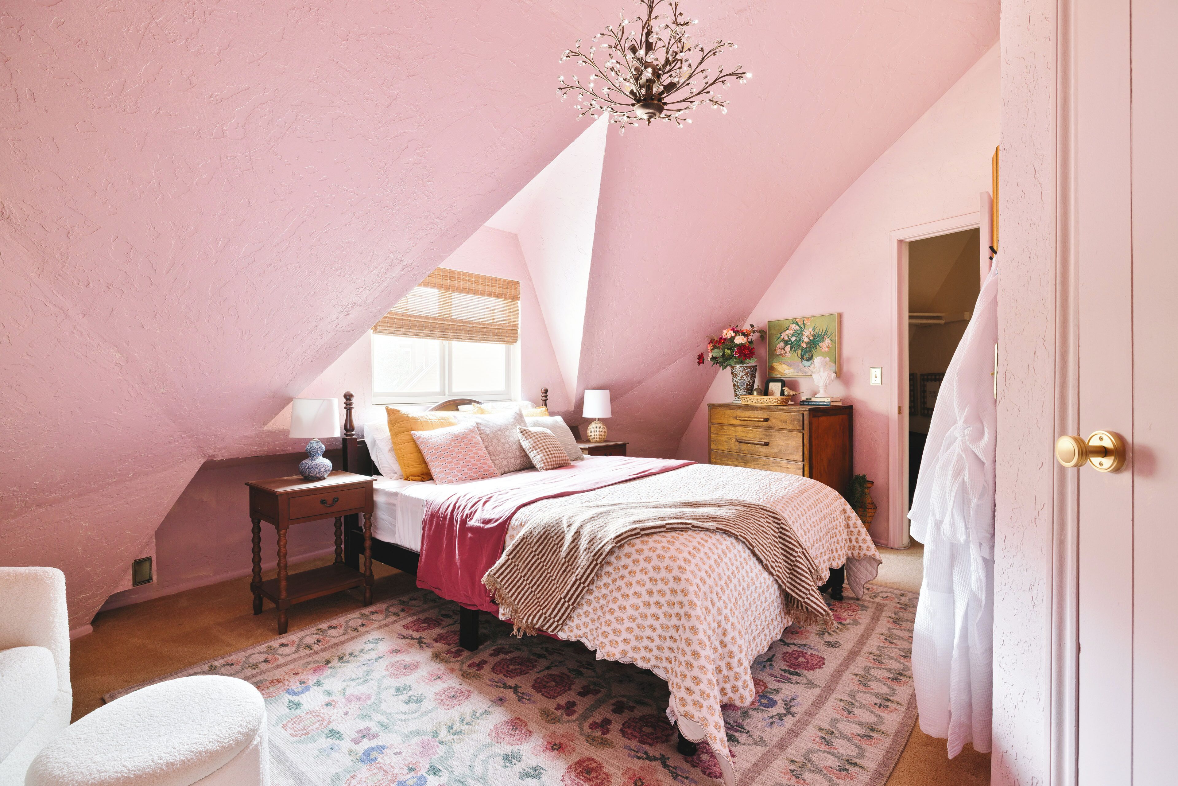 Une chambre avec un plafond en pente, un lit recouvert d’une couvre-lit à motifs floraux, une table de chevet avec une lampe et une commode avec un miroir.
