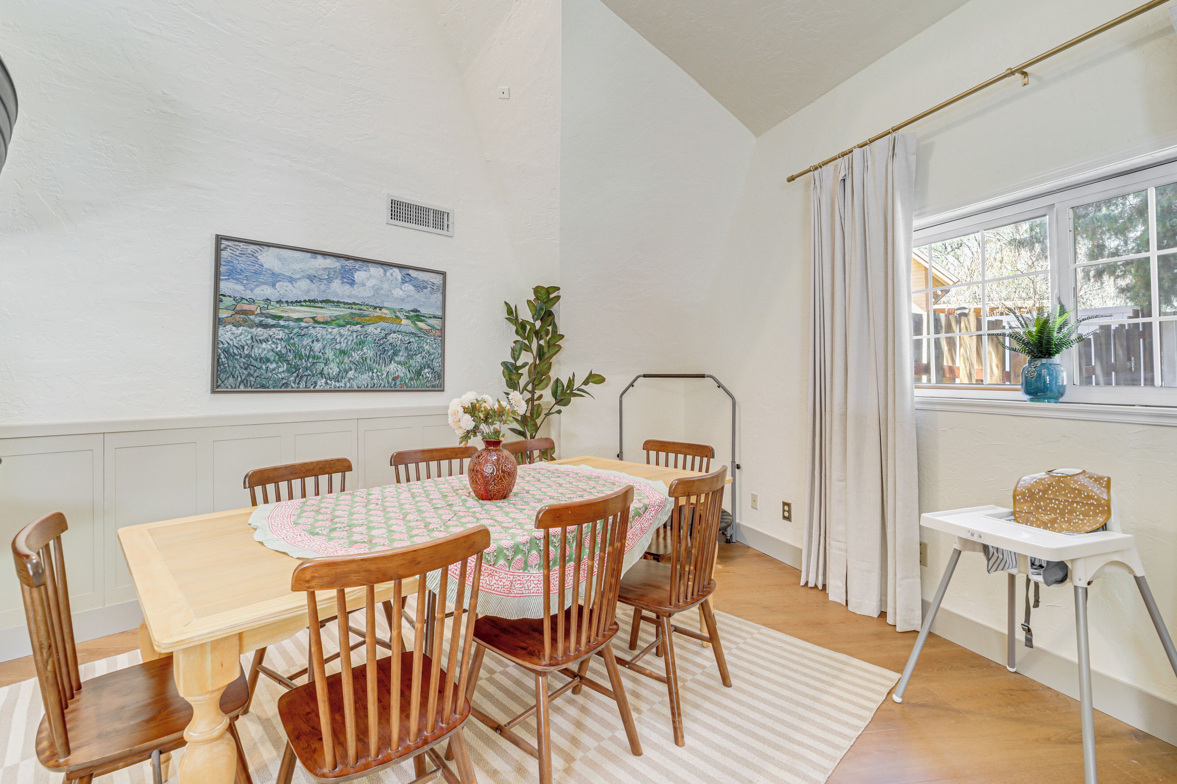 A dining area with a wooden table, chairs, a vase with flowers, and a painting on the wall.