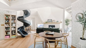 Een ruime woonkamer met houten vloer, een spiltrap, een open haard en een eethoek met een ronde tafel en stoelen.