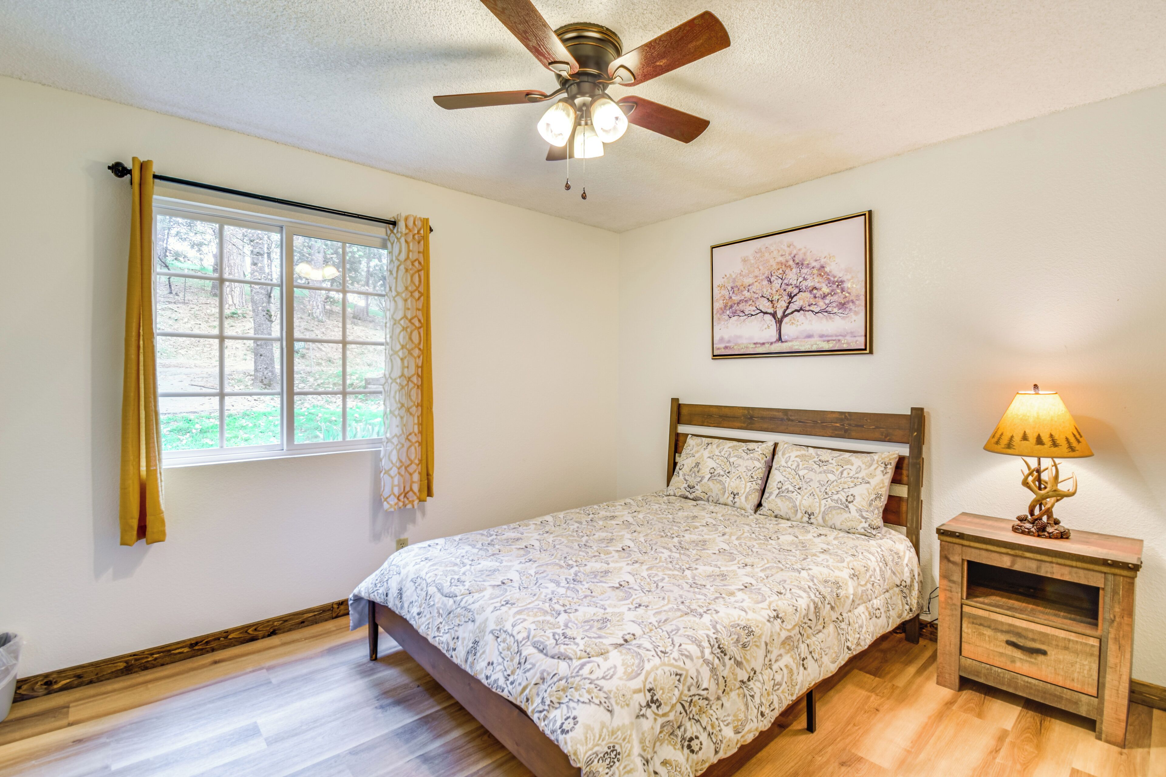 A bedroom with a bed, a ceiling fan, a window with curtains, a bedside lamp, and a framed picture on the wall.