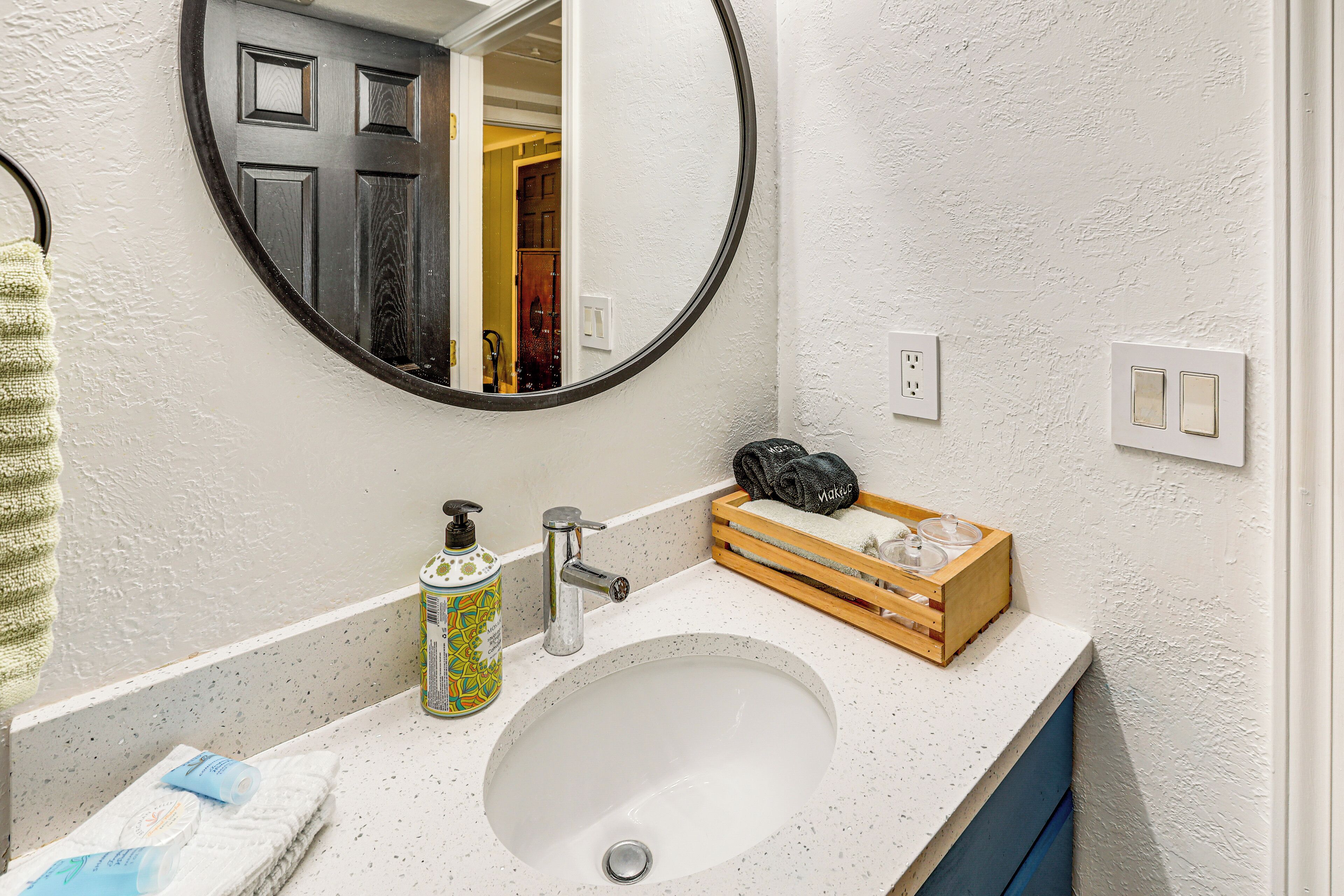A bathroom with a white sink, a round mirror, a wooden box with toiletries, and a towel hanging on the wall.