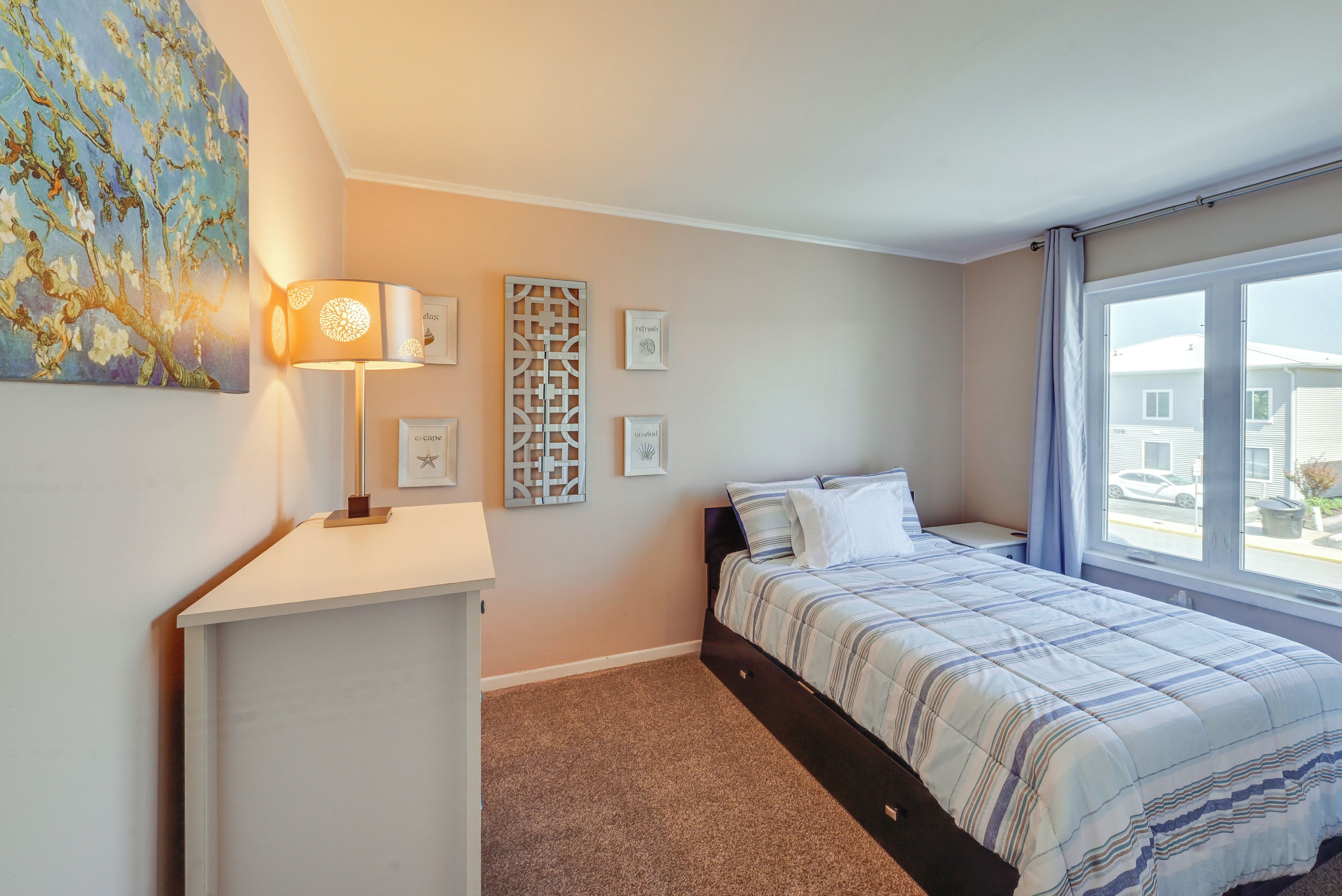 A bedroom with a bed, a desk, a lamp, and a window with a view of a house.