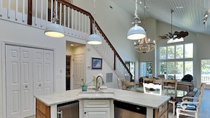 A spacious kitchen with a central island, white cabinetry, and a dishwasher. A staircase leads to a loft area with a dining space. - The Pack House | Waterfront Home, Private Dock (Topsail Beach)