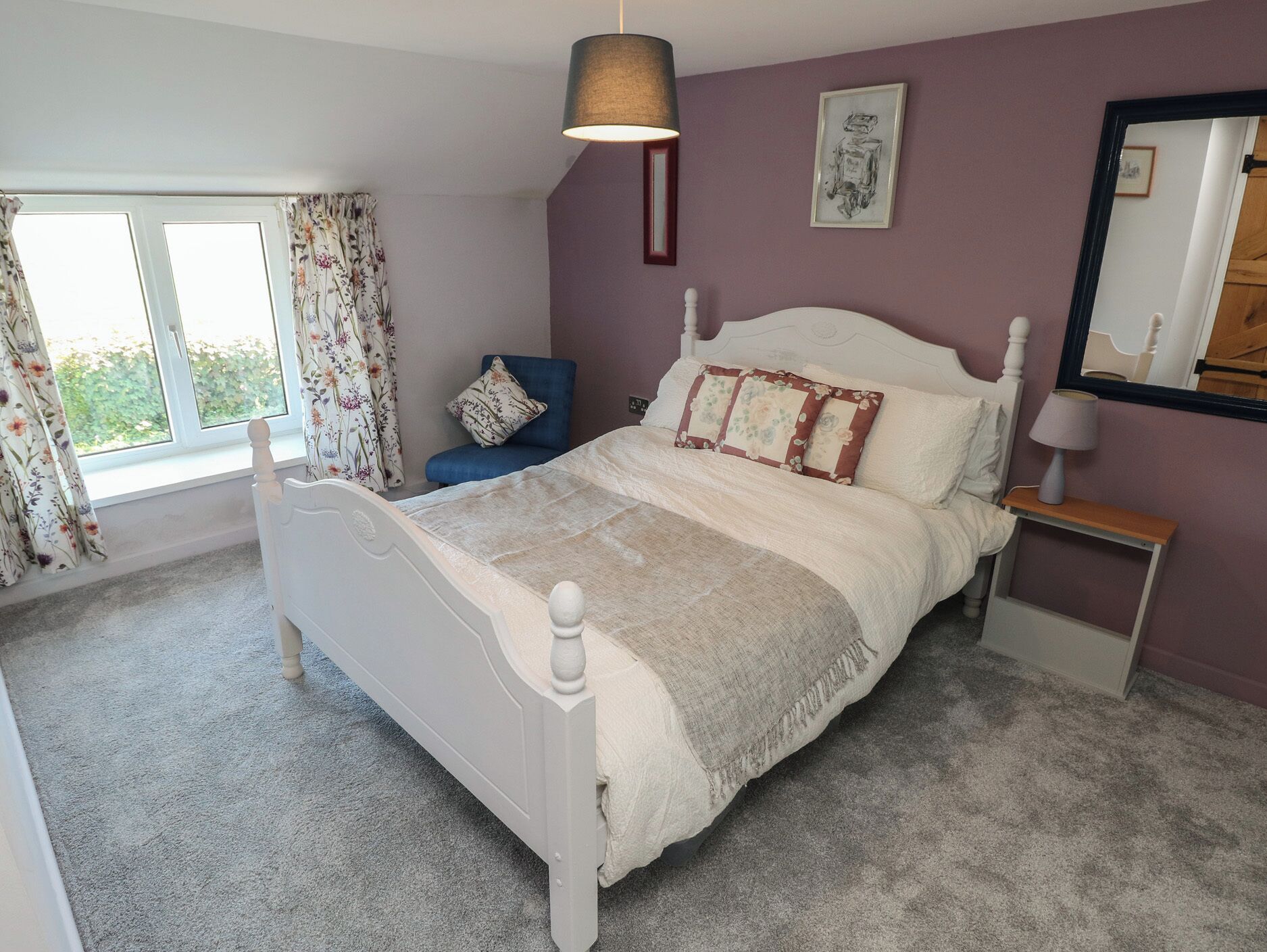 Une chambre avec un lit blanc, des rideaux à motifs floraux, une chaise bleue, une table de chevet avec une lampe et un miroir au mur.