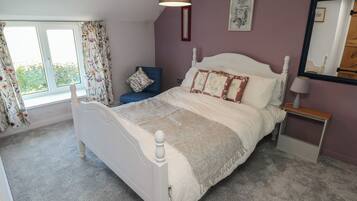 Une chambre avec un lit blanc, des rideaux à motifs floraux, une chaise bleue, une table de chevet avec une lampe et un miroir au mur.