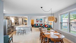 Uma cozinha moderna com uma ilha central, uma sala de jantar com mesa e cadeiras de madeira, e uma sala de estar com sofá e almofadas decorativas.