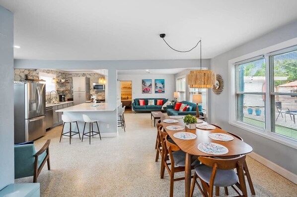 A modern kitchen with a central island, a dining area with a wooden table and chairs, and a living area with a sofa and decorative pillows.