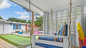 Uma área de estar coberta ao ar livre com um balanço, uma piscina e um edifício branco ao fundo.