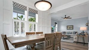 A spacious dining area with a wooden table and chairs, large windows with white shutters, a ceiling fan, and a living area with a sofa and lamp. - Downtown St Augustine Steps to Ice Plant Upscale (St. Augustine)