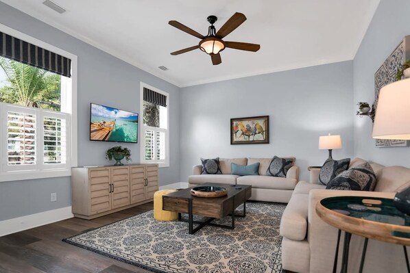 A living room with a sofa, coffee table, and a ceiling fan. There are windows with blinds, a TV, and a painting on the wall. - Downtown St Augustine Steps to Ice Plant Upscale (St. Augustine)