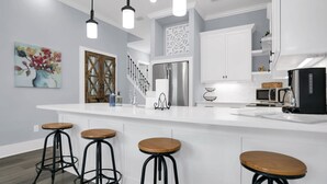 A modern kitchen with white cabinetry, a stainless steel refrigerator, a microwave, and a coffee maker. There are three wooden stools at the island. - Downtown St Augustine Steps to Ice Plant Upscale (St. Augustine)