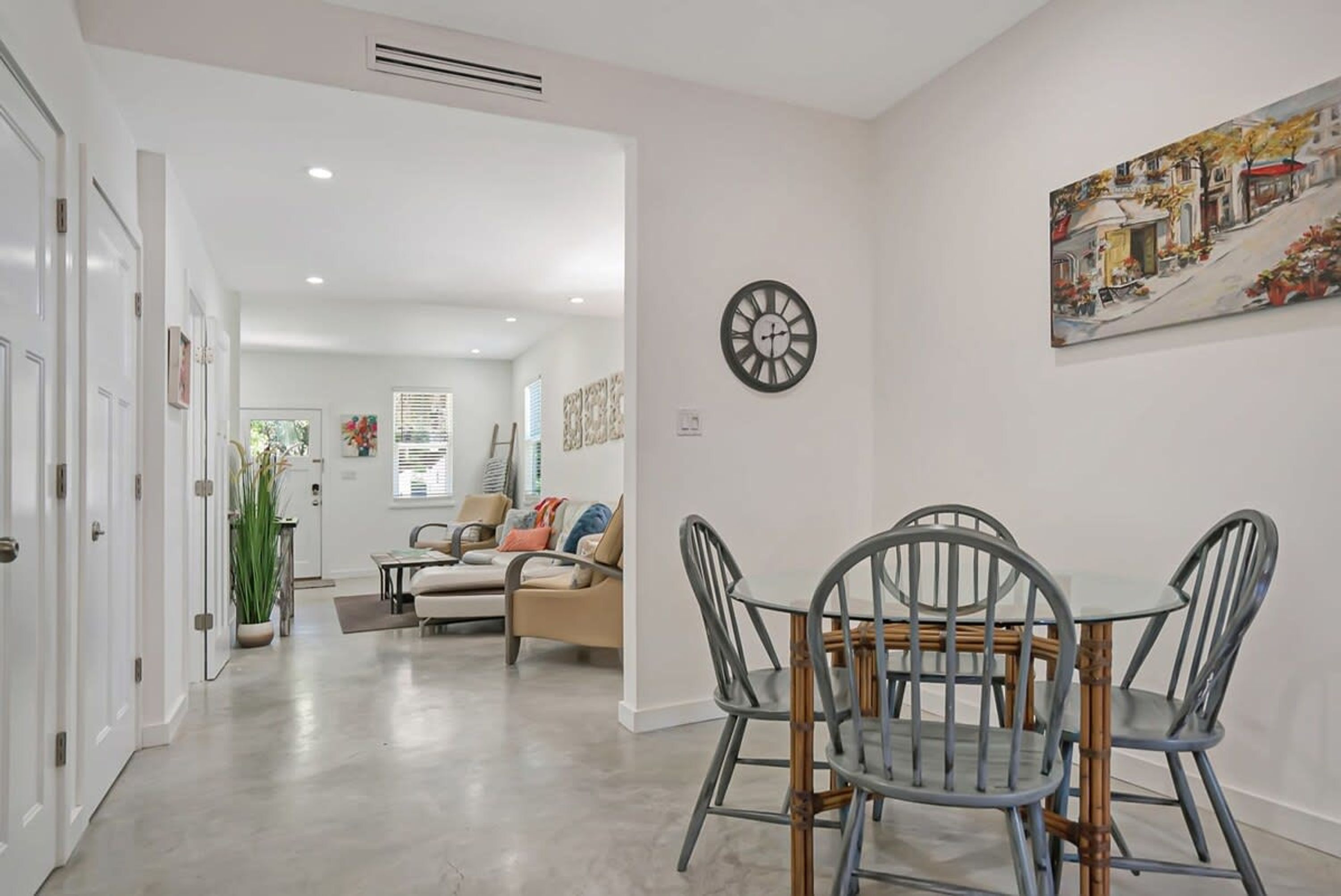 A dining area with a round table and four chairs, a clock on the wall, and a painting depicting a street scene.