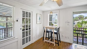 A room with a wooden floor, white walls, a ceiling fan, a dining table with chairs, and large windows with blinds. - Marvelous Mermaid Beach Bungalow Dog-friendly (St. Augustine)