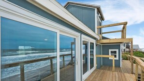 Een gebouw aan de kust met grote glasramen, een houten terras en uitzicht op de oceaan.