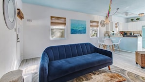 Un salon moderne avec un canapé bleu, un coin repas avec une table et des chaises blanches, et une cuisine avec des meubles blancs et un plan de travail bleu.