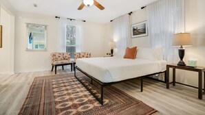 Quarto com cama, ventoinha no teto, mesa de cabeceira, abajur, cadeira, tapete e janela com cortinas.