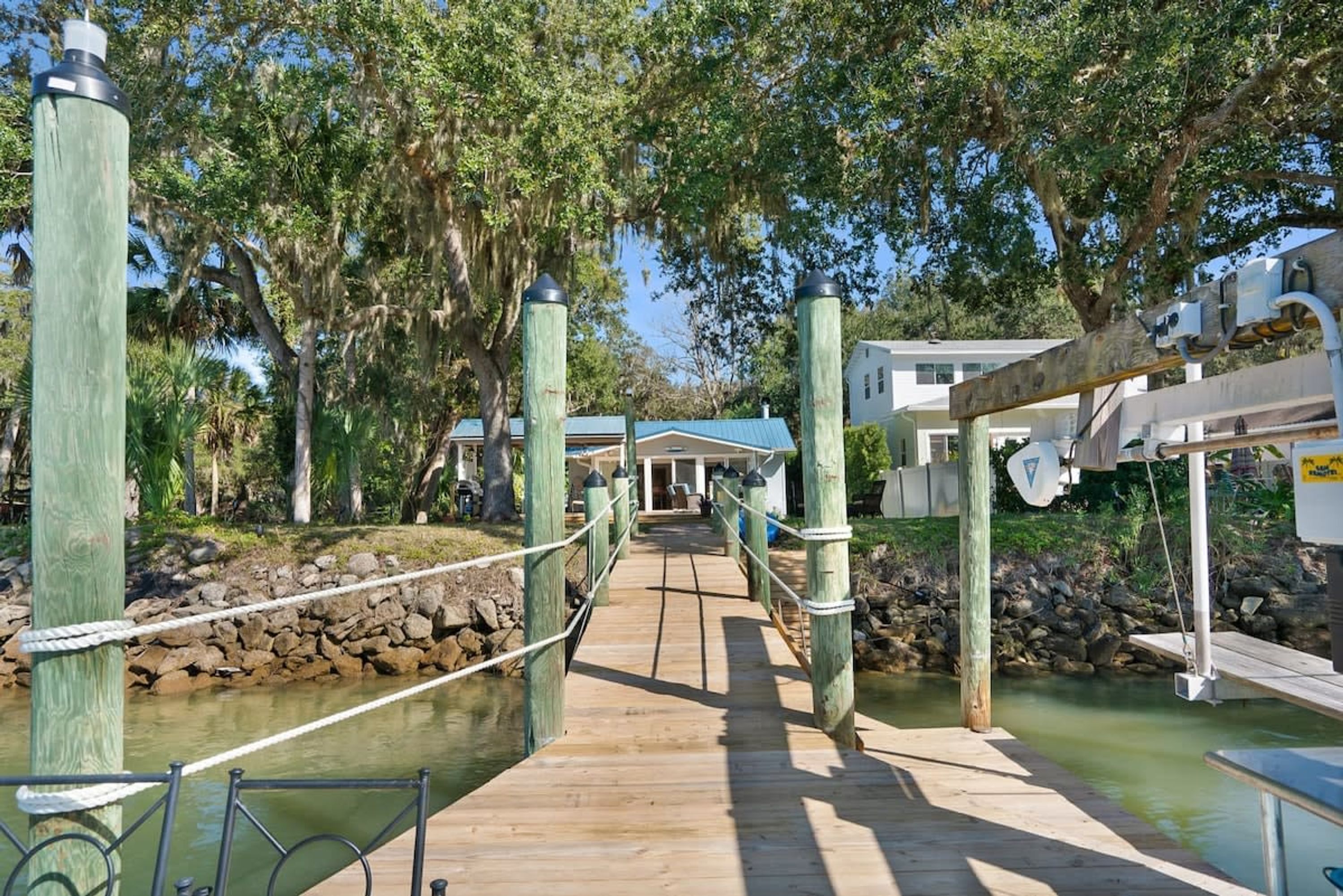 A wooden bridge over a calm water body, surrounded by lush greenery and a building with a blue roof in the background.
