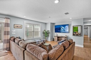 Een moderne woonkamer met een hoekbank, een flatscreen televisie en een groot raam met zonwering.