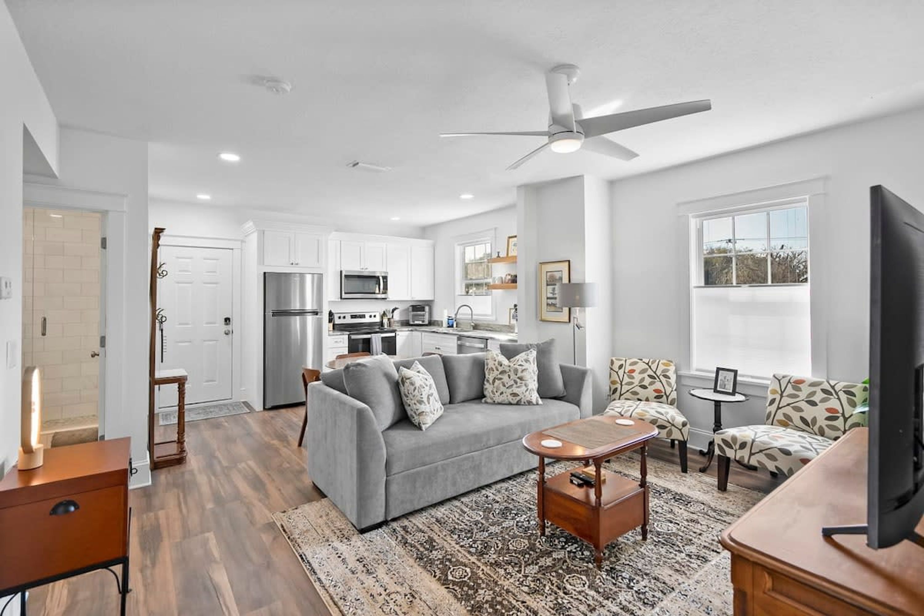 A modern living room with a grey sofa, a coffee table, and patterned chairs. A TV is on a wooden stand. The room has a ceiling fan, a kitchen in the background, and a large window with a view. - Free Parking Big Patio Next to Restaurants Bars (St. Augustine)