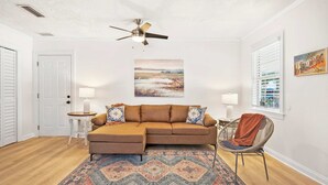 A living room with a brown sectional sofa, a patterned rug, a ceiling fan, a painting, and a window with blinds. - Just North of Historic St Aug Patio Garage BBQ (St. Augustine)