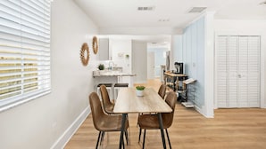 A modern kitchen with white cabinets, a wooden dining table with four chairs, and a view of the adjacent dining area. - Just North of Historic St Aug Patio Garage BBQ (St. Augustine)