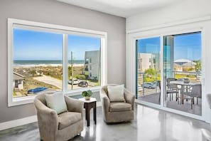 A spacious room with large windows offering a view of the beach and ocean. There are two armchairs and a small table with a plant. - Ocean Vista Beach House Pool Spa Steps To Sand (St. Augustine)
