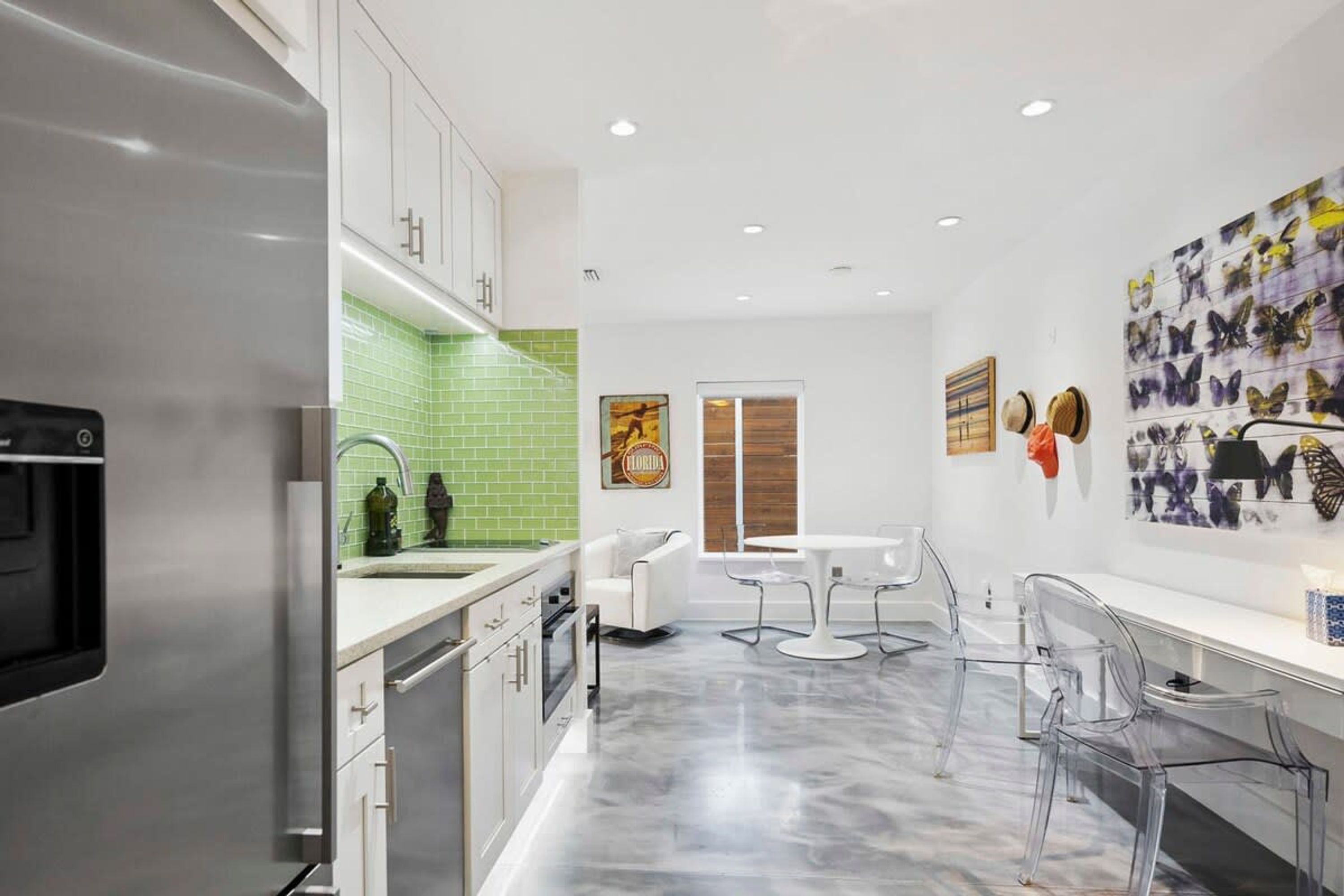 A modern kitchen with white cabinets, a green backsplash, and a marble floor. There is a dining area with a white table and chairs.