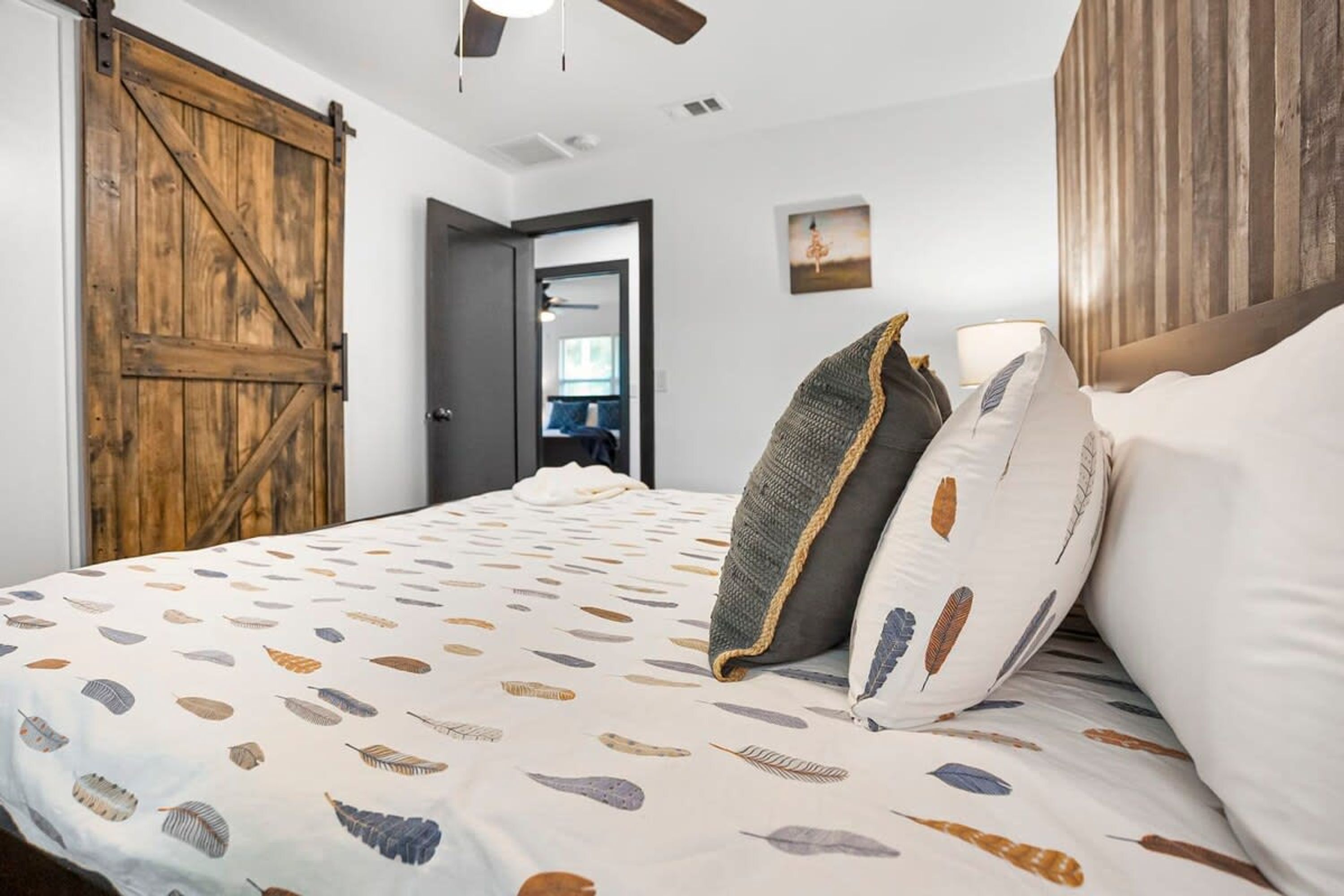 A bedroom with a large wooden door, a bed with patterned bedding, and a view of a room with a window and a chair.