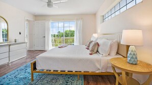 A bedroom with a bed, a bedside table with a lamp, a dresser, a ceiling fan, a large window, and a balcony.