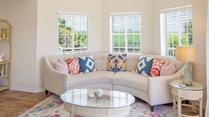 A spacious living room with a curved white sofa, a glass coffee table, and a colorful rug. There are three windows with white shutters, a ceiling fan, and a small side table with a lamp.