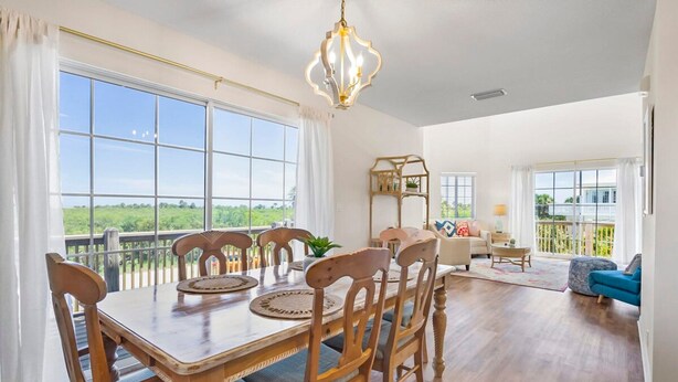 A spacious dining area with a wooden table and chairs, large windows offering a view of greenery, and a balcony with a railing.
