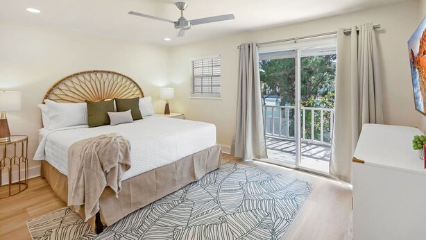 Ein Schlafzimmer mit Bett, Deckenventilator, Fenster mit Vorhängen und Blick ins Freie.