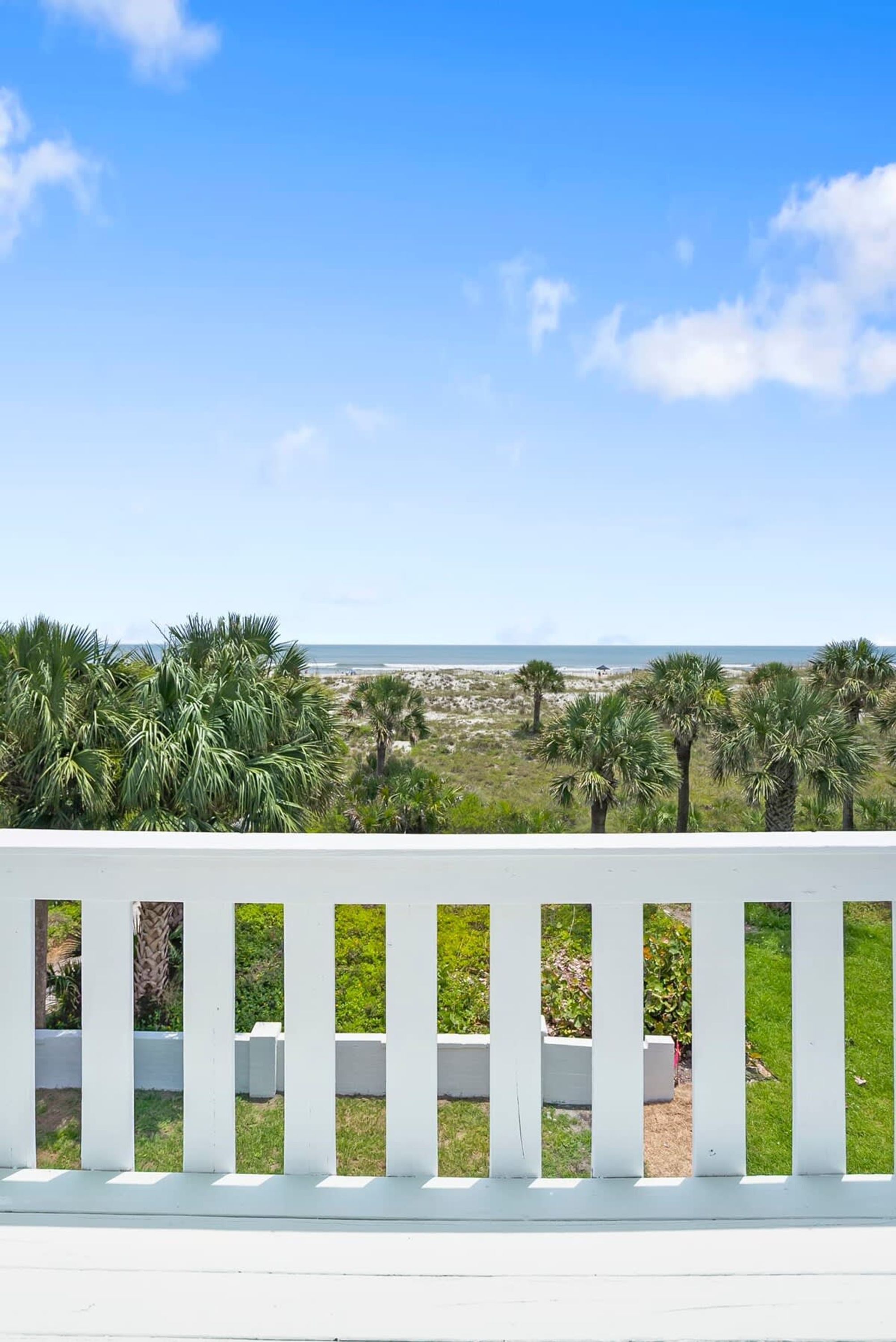 A balcony with a white railing overlooking a beach with palm trees and a clear sky.