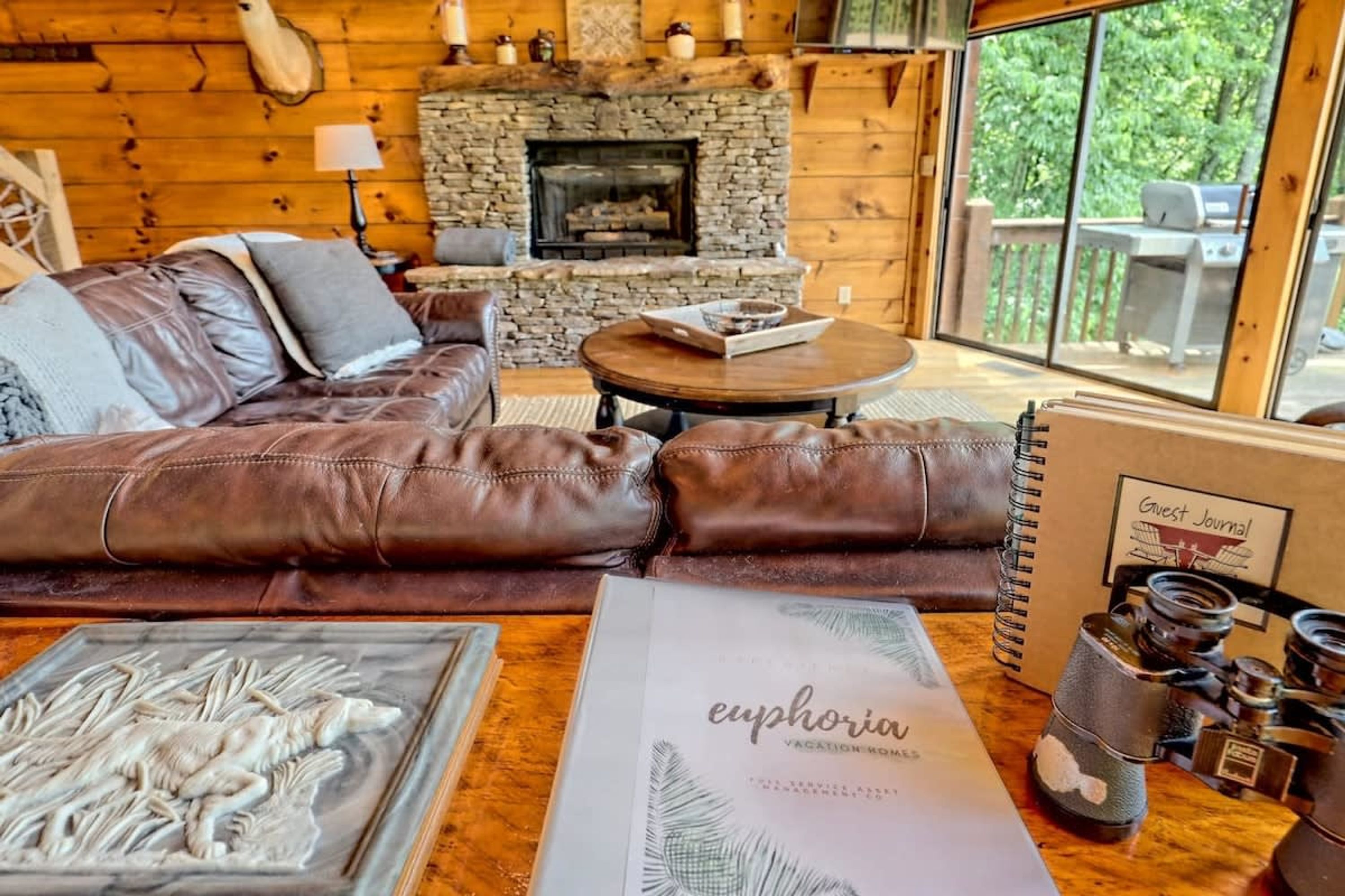 A cozy cabin interior with a stone fireplace, leather sofa, and a large window overlooking greenery.