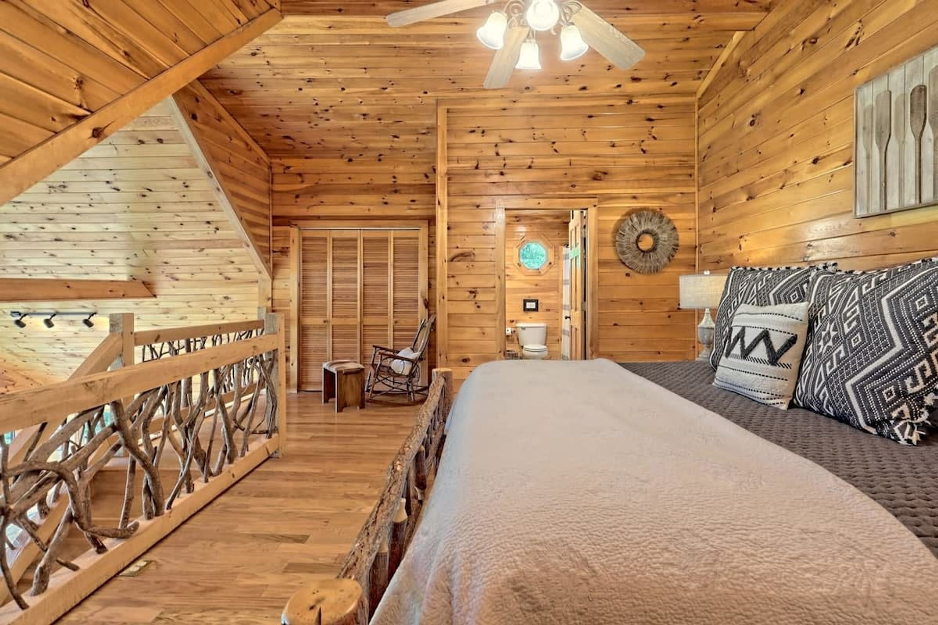 A wooden cabin interior with a bed, a chair, a small table, and a ceiling fan.