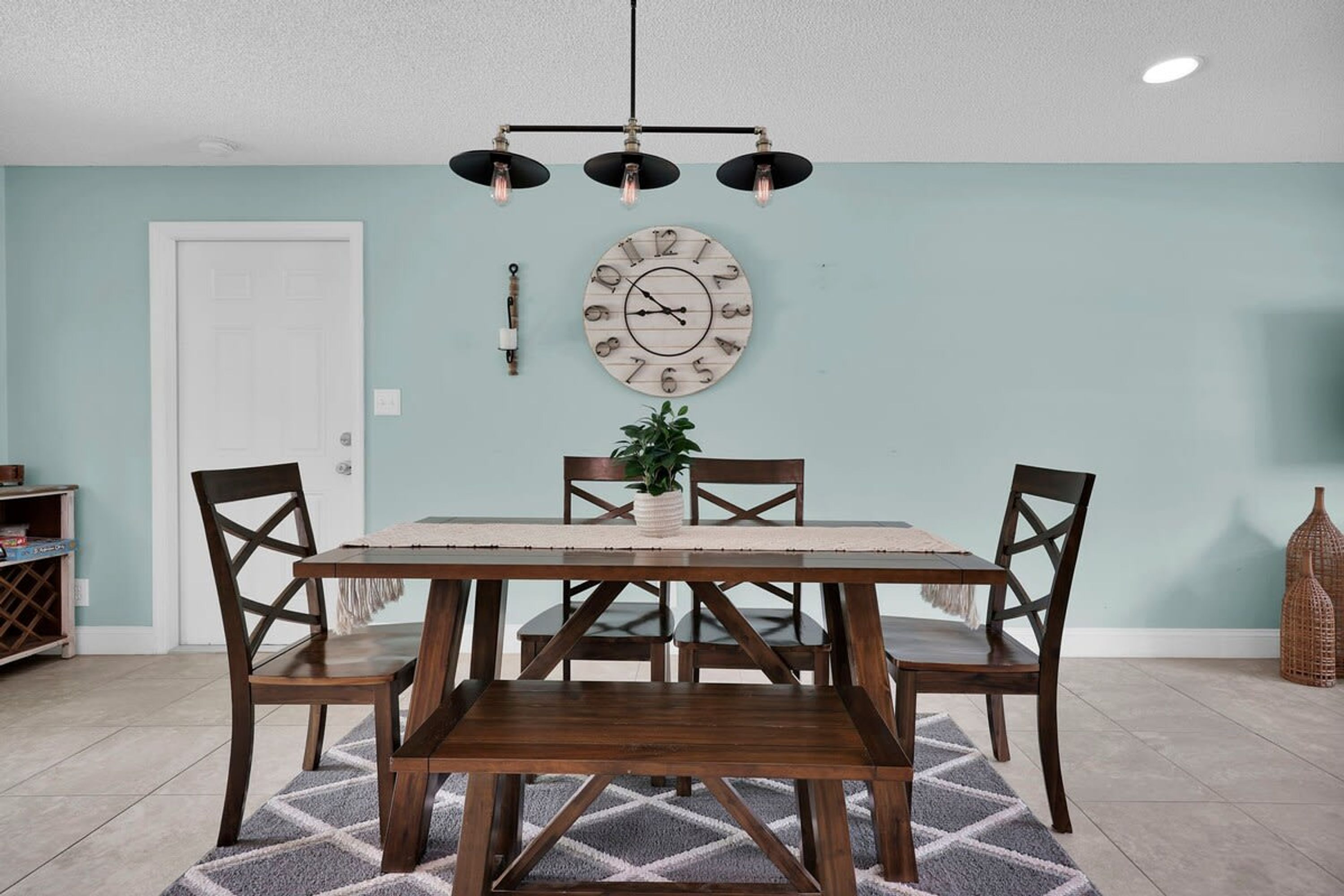 A dining area with a wooden table and chairs, a wall clock, and a hanging light fixture.