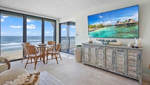 A balcony with a view of the ocean, a dining area with wicker chairs, and a large flat-screen TV on a rustic wooden cabinet.