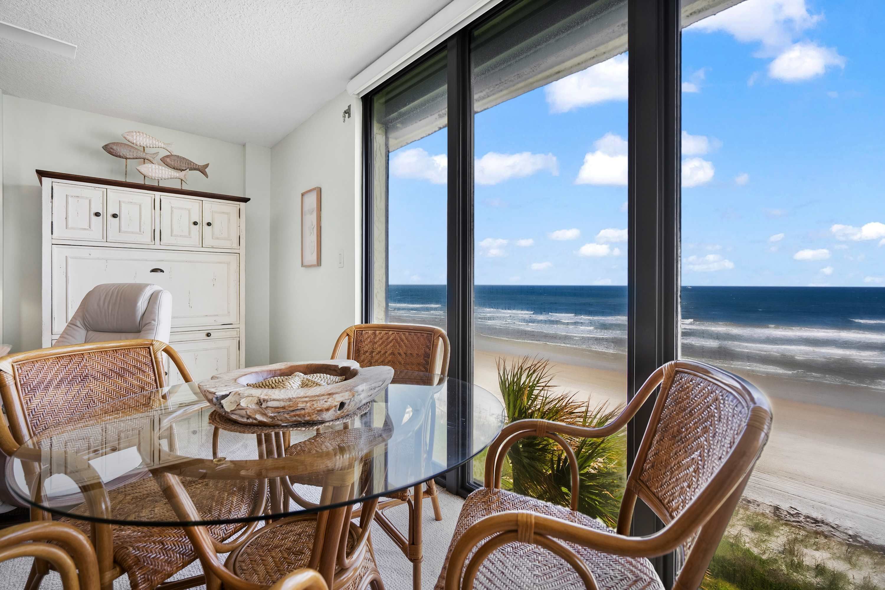 A room with a glass table, wicker chairs, a white cabinet, and a view of the ocean.