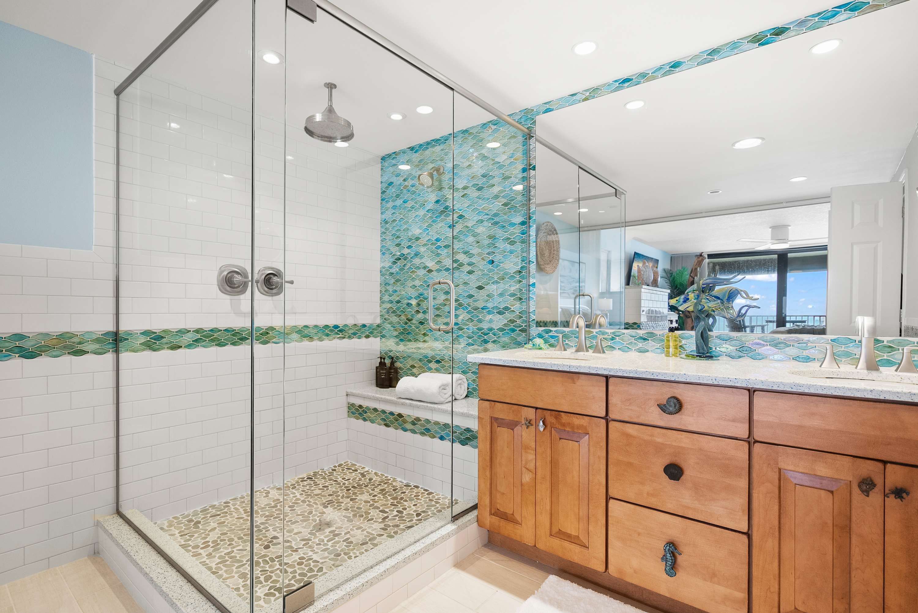 A modern bathroom with a glass-enclosed shower, a wooden vanity with multiple drawers, and a tiled backsplash with a green and blue pattern.