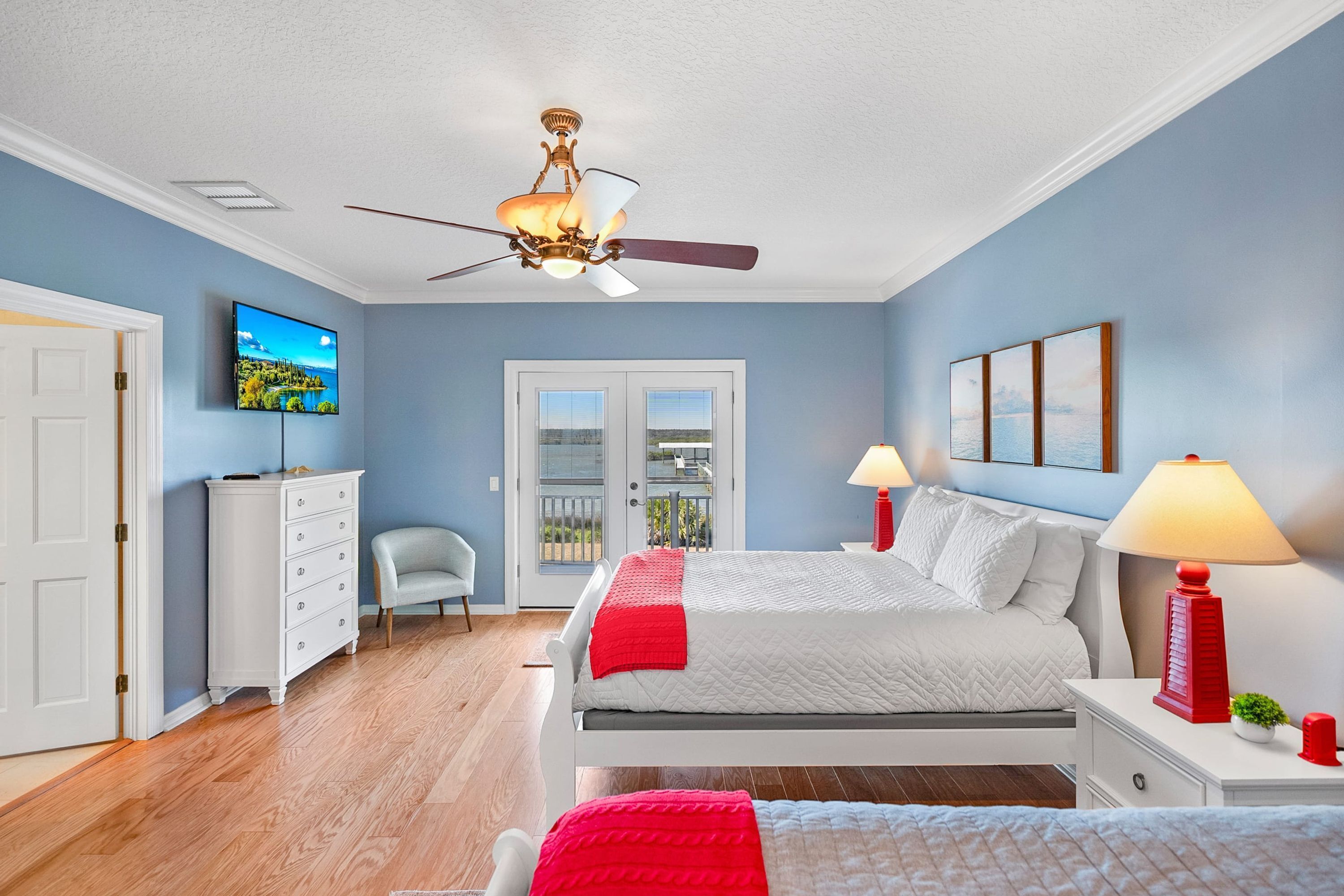 Ein Schlafzimmer mit Bett, Deckenventilator, Fernseher, Kommode, Stuhl und Balkontür.