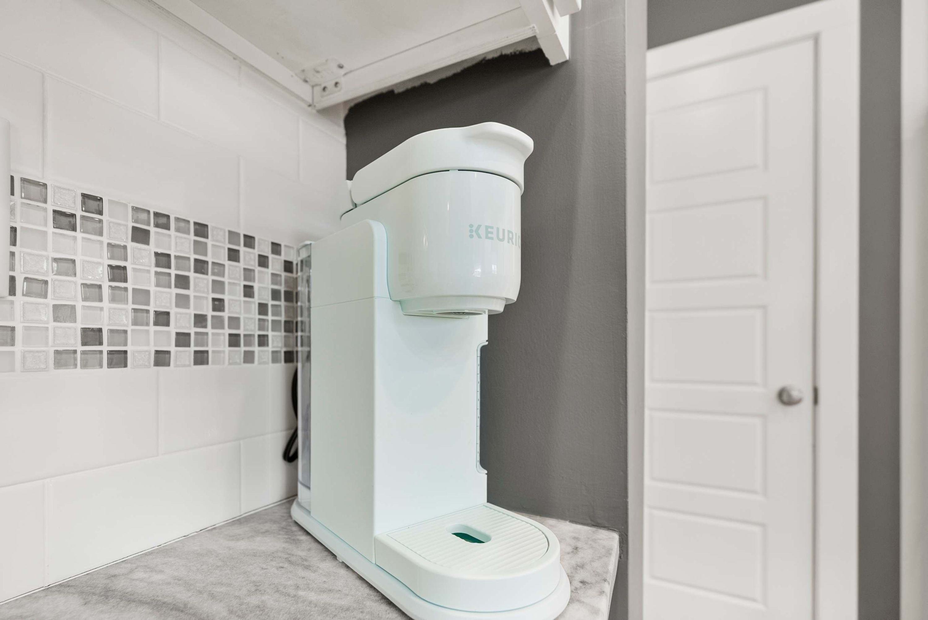 Une machine à café Keurig blanche est posée sur un comptoir en marbre dans une salle de bain aux murs recouverts de carreaux blancs et dont la porte est également blanche.
