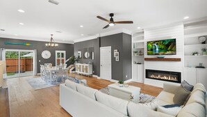A modern living room with a fireplace, a television, and a dining area with a view of the backyard. - Magnolia Ave Downtown By Beach Kids Playset (St. Augustine)