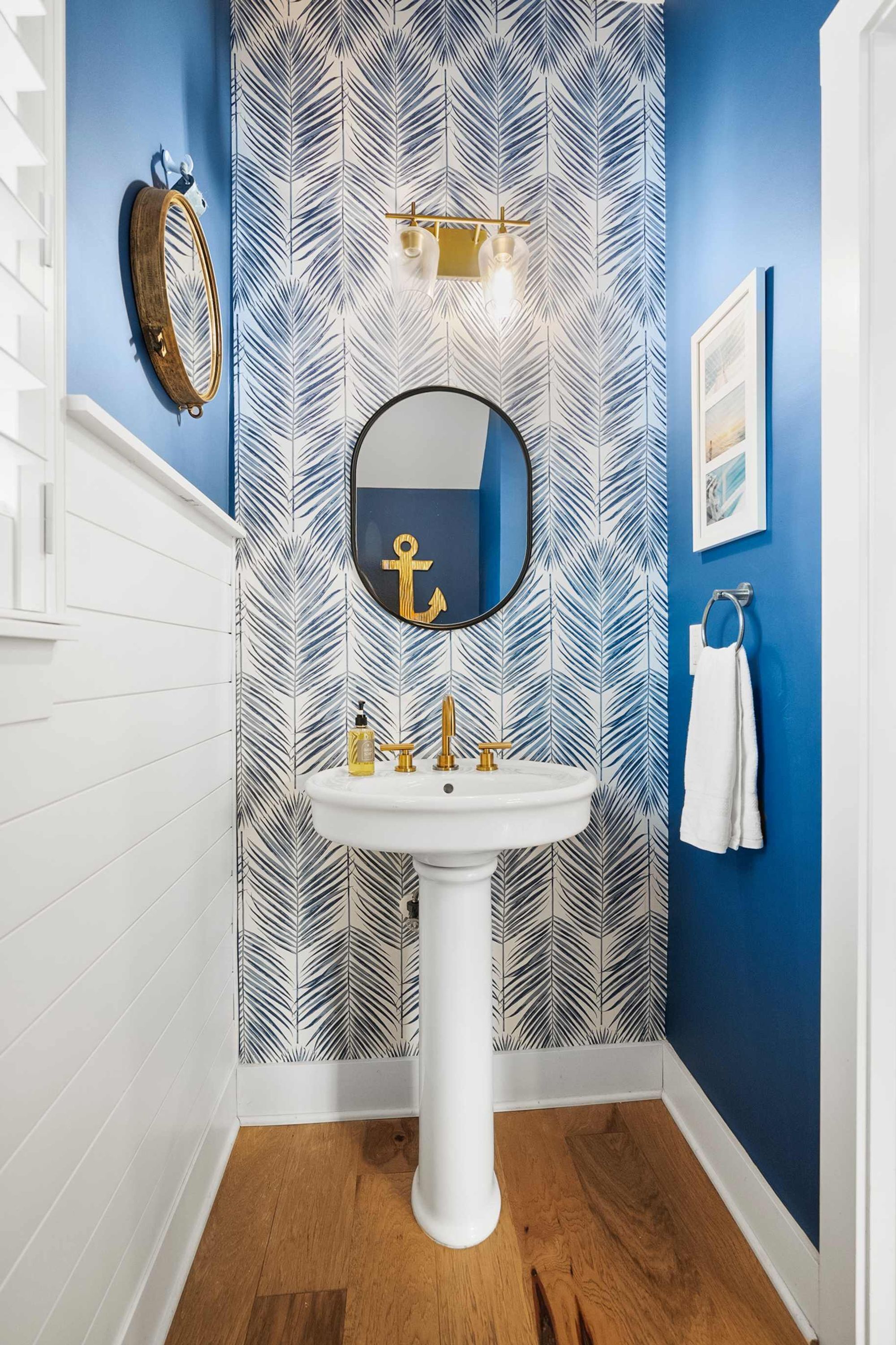 Un baño con un lavamanos pedestal blanco, un espejo redondo con diseño de ancla y papel tapiz estampado con hojas.