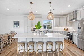 A modern kitchen with white cabinets, a large island with bar stools, stainless steel appliances, and hanging pendant lights. - Your Summer Haven Pool Games Near Beach (St. Augustine)