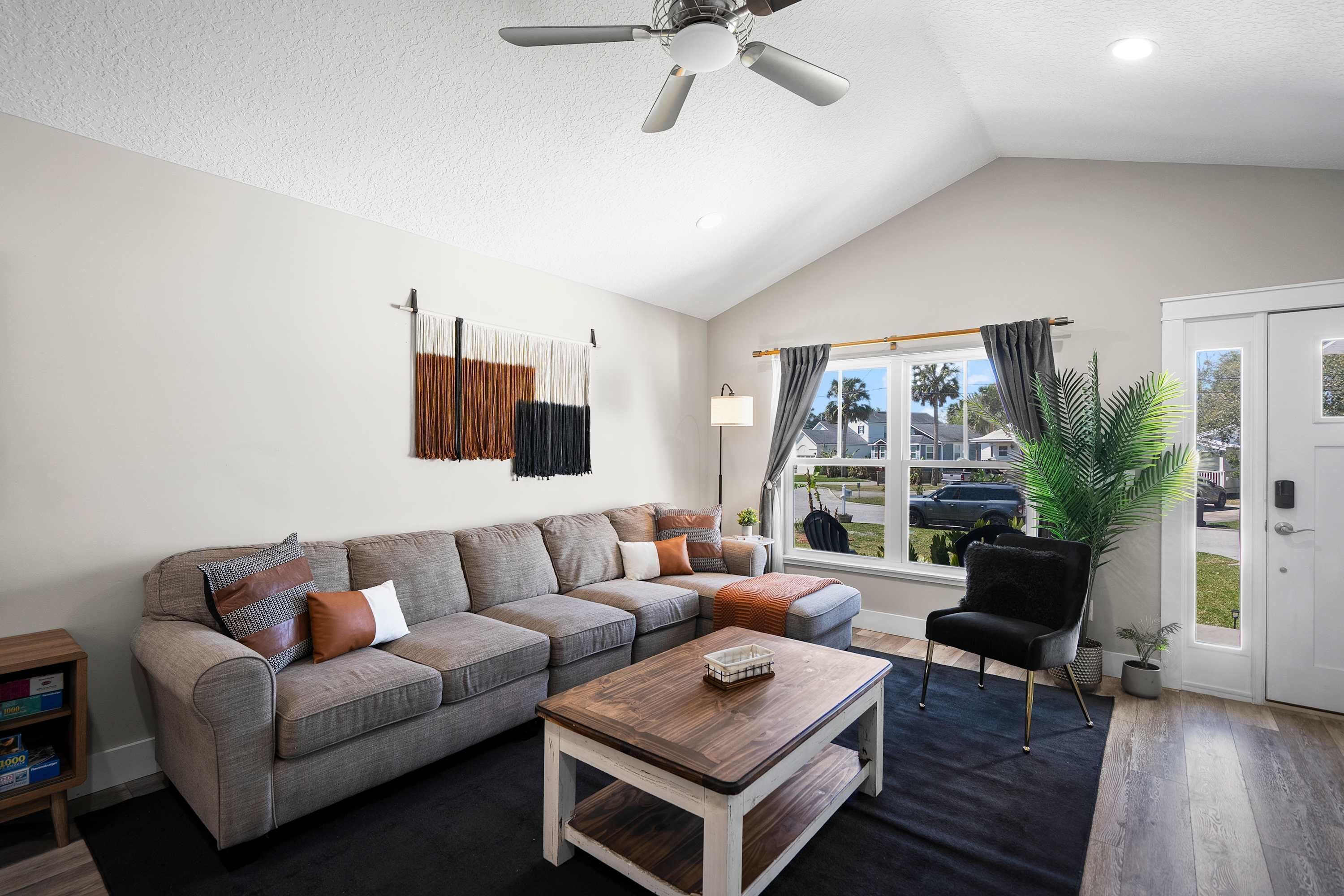 A living room with a grey sectional sofa, a wooden coffee table, a black armchair, and a large window with curtains.